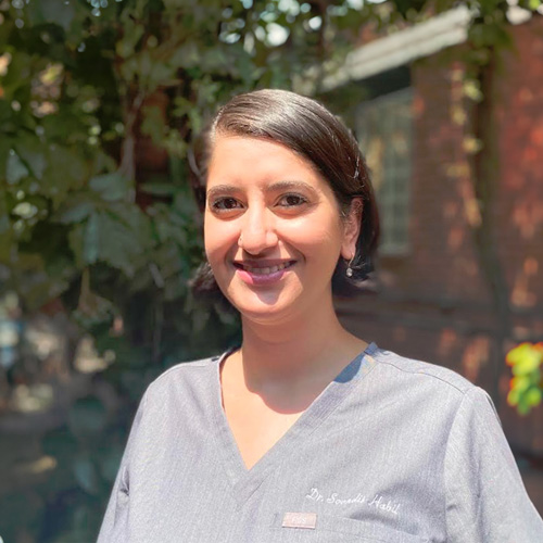 A smiling woman wearing scrubs stands outdoors with trees behind her.
