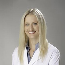 The image shows a woman wearing a white lab coat, standing against a light-colored background, smiling at the camera.