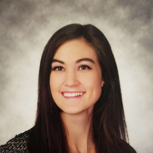 The image shows a woman with long dark hair smiling at the camera, wearing a black top with a necklace, standing against a blurred background that appears to be a school photo backdrop.