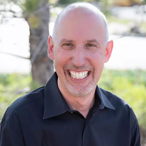 The image shows a man smiling at the camera he appears to be indoors with a blurred background that suggests an outdoor setting, possibly a patio or garden.