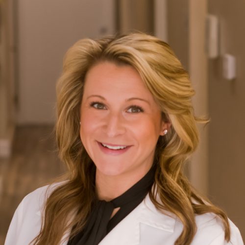 A smiling woman wearing a white coat stands confidently in an office setting, with a warm and welcoming expression.