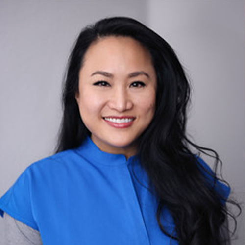 The image shows a person with long hair smiling at the camera. They are wearing a blue jacket over a light-colored top.