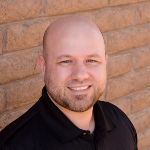The image shows a man with a bald head smiling at the camera, wearing a black shirt and standing in front of a brick wall.