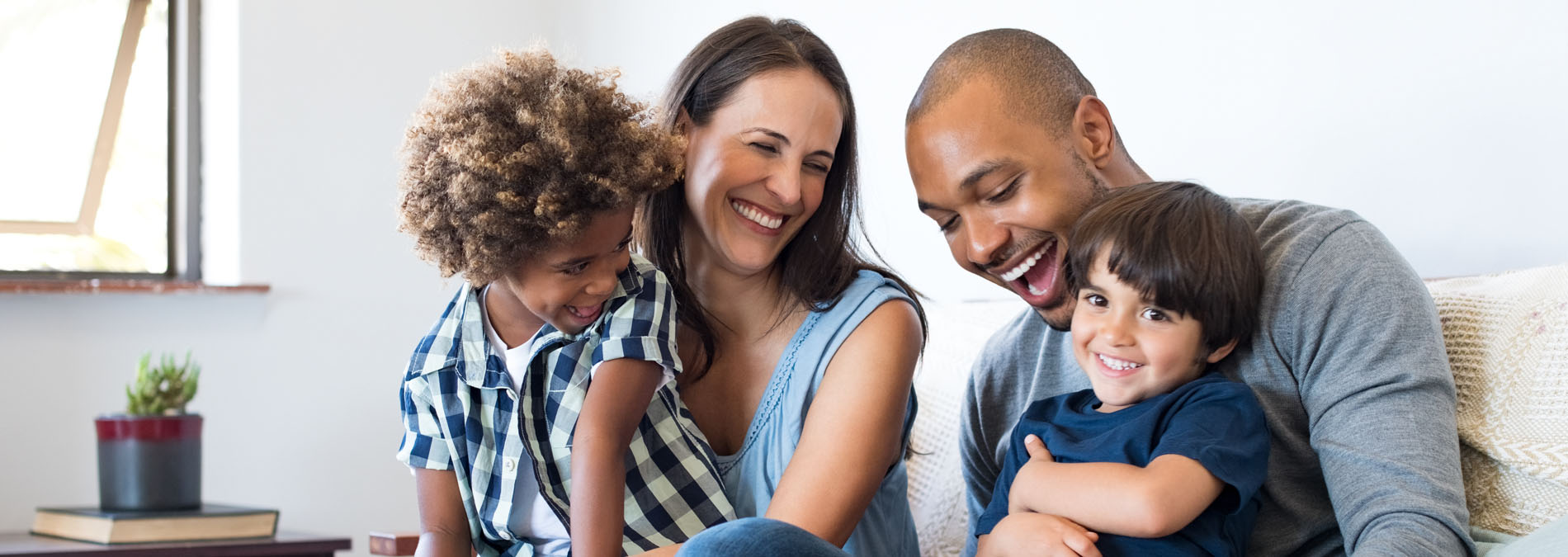 The image depicts a family of four, with two adults and two children, sitting together on a couch, smiling at the camera, with a man holding a young child on his lap.