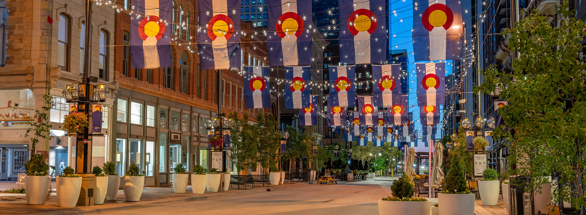 The image shows a festively decorated street at night with illuminated flags and string lights, featuring a variety of colors and patterns, set against a backdrop of urban buildings and a clear sky.