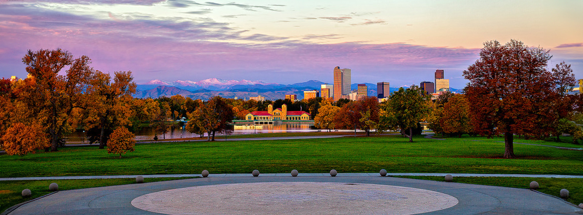 The image shows a circular stone structure, possibly a memorial or monument, with a panoramic view of a city skyline at sunset or sunrise, featuring warm colors in the sky and a clear horizon line.