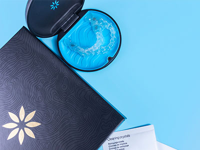 The image shows a closed box with a patterned design on its lid placed next to an open box containing a blue item, possibly a product, with a small amount of water visible inside the box, against a blue background.