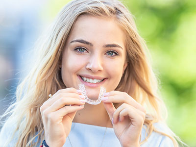 The image shows a young woman with a radiant smile holding up her teeth, posing for a photo with her mouth open wide, displaying her dental braces.