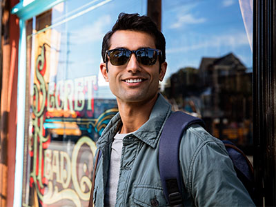 The image features a man standing outside a storefront with sunglasses on, wearing a backpack and a jacket, appearing relaxed and posing for the photo.