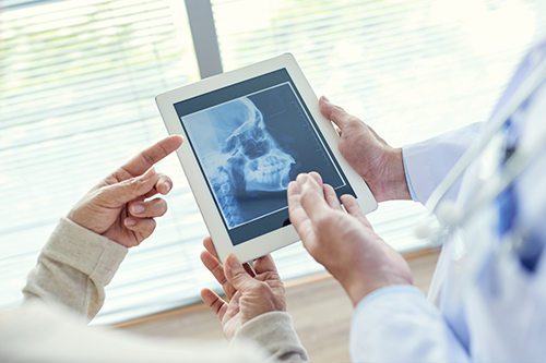 The image depicts two individuals using a tablet computer to view an X-ray, with a medical professional holding the device and pointing at it while standing next to another person who appears to be seated and engaged in the discussion.