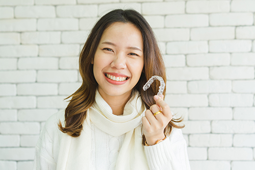 A smiling woman holding up a sign with a smiley face on it, standing against a brick wall.