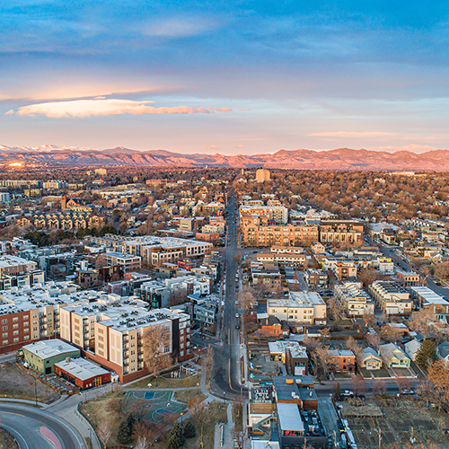 The image shows an urban landscape during sunset, featuring a city skyline with buildings, streets, and a clear view of mountains in the background, under a partly cloudy sky.