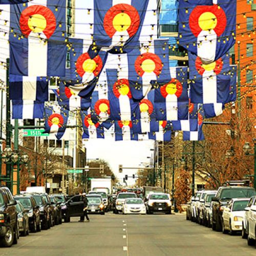The image shows a city street lined with cars, decorated with banners featuring the Colorado state flag, and festooned with flags and lights.