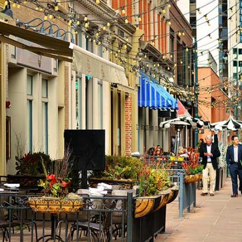 The image depicts a vibrant city street lined with outdoor dining restaurants and shops, featuring a variety of architectural styles and decorative elements such as string lights, planters, and awnings.