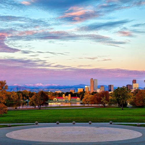 The image depicts a serene cityscape at sunset with a prominent circular fountain in the foreground, a large city skyline in the background, and a clear sky above.