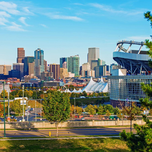 The image shows a city skyline with a prominent stadium in the foreground, featuring a large dome structure and various other buildings, under a clear blue sky.