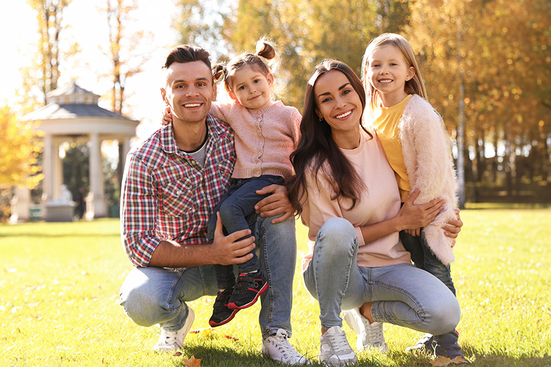 The image depicts a family of four, consisting of a man, woman, and two children, posing for a photograph outdoors during daylight hours with trees and grass visible in the background.