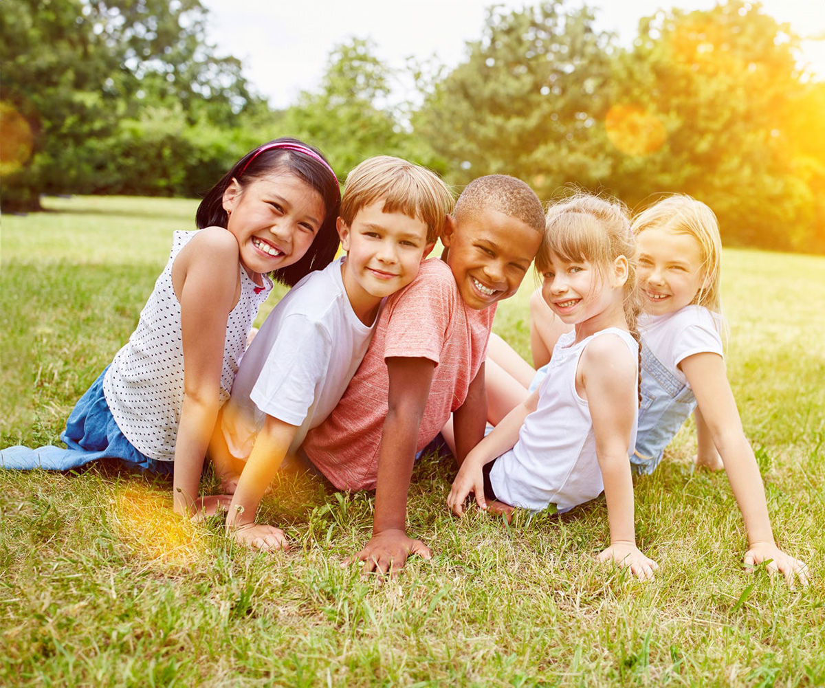 This is a color photograph featuring four children and an adult posing together outdoors on grass. They appear to be smiling and enjoying themselves, with the adult standing behind them. The lighting suggests it might be late afternoon or early evening.