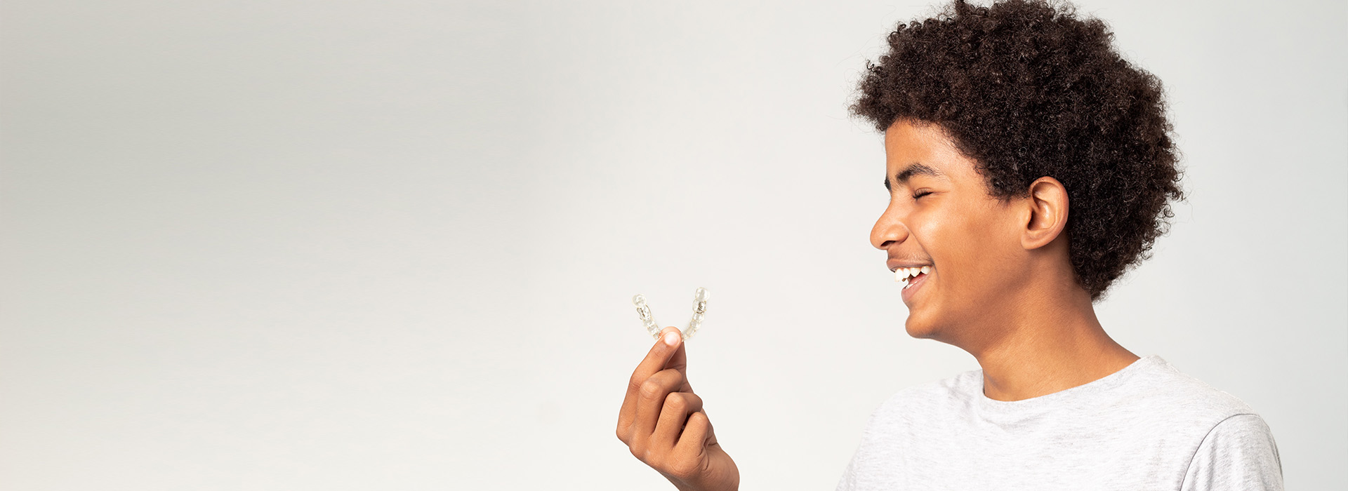 The image shows a young person with curly hair smiling at the camera, holding a small object in their hand, against a plain background.