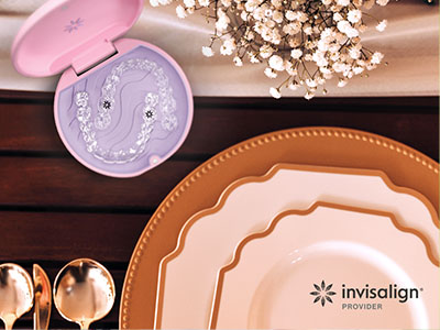 The image shows a dining table set with plates, cutlery, and a cake stand, featuring a pink cake box with an illustration of a mermaid on top, placed on a wooden surface with decorative items and a  Invisalign  logo visible in the bottom right corner.
