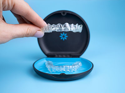 The image shows a person s hand holding an open dental impression tray containing a set of clear aligners, with the tray placed on a blue object that resembles a case, and the background is a solid color.