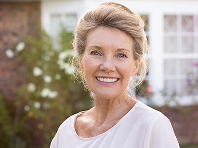 A woman with blond hair wearing a white top stands outside a house with a smile on her face.