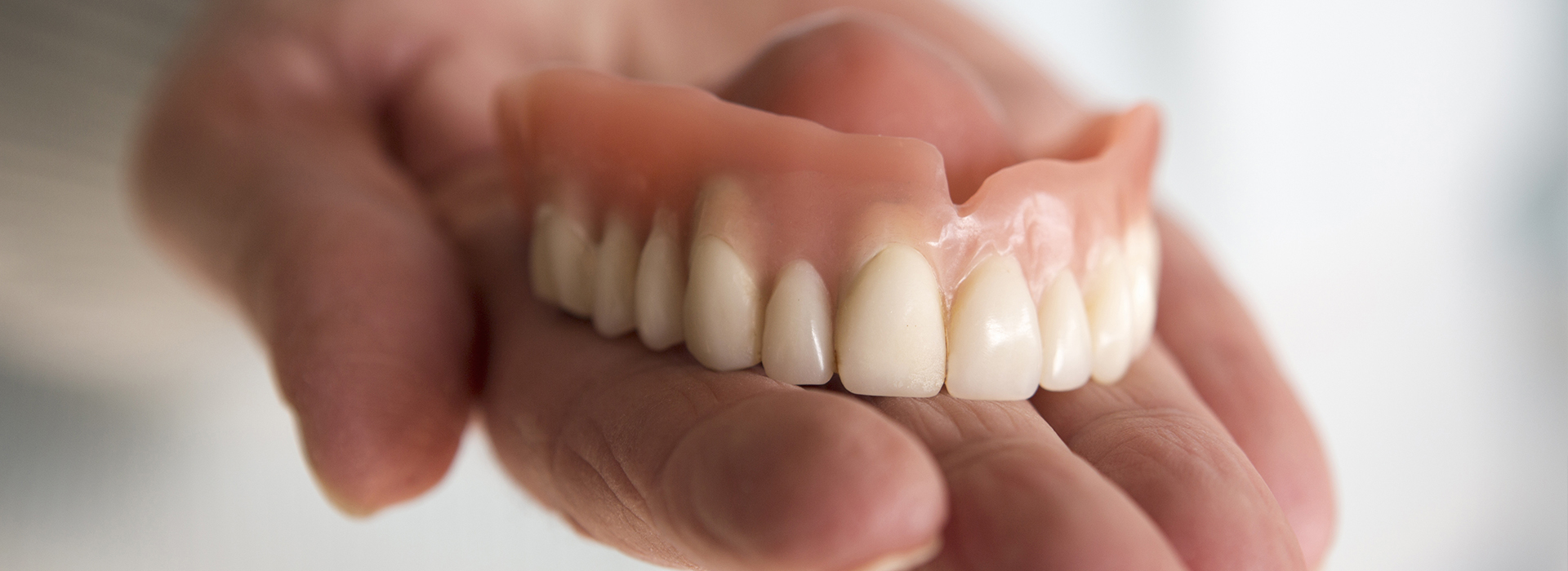 The image features a hand holding a set of dentures against a blurred background.