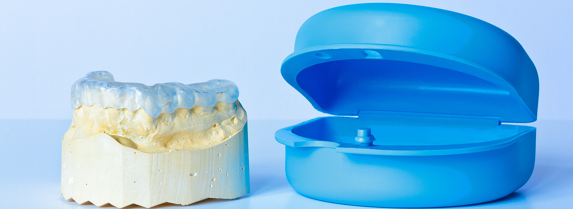 The image shows two dental implants, one in a blue plastic container and the other in a yellowish-brown material, against a white background.
