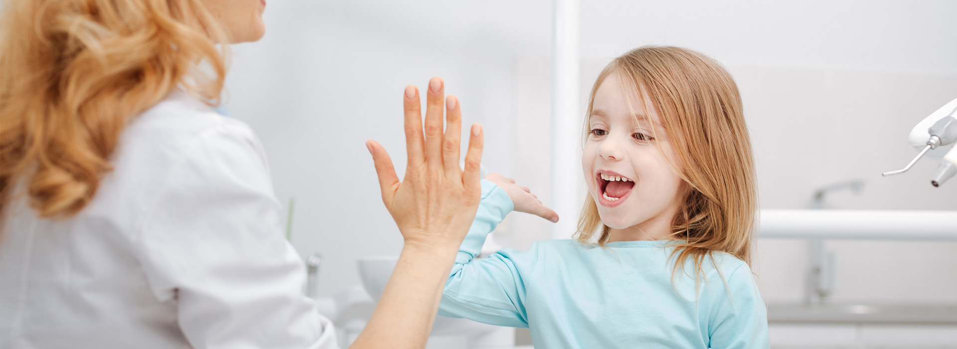 The image shows a young girl waving at a woman who appears to be interacting with her from behind a counter, likely in a dental or medical setting, as indicated by the presence of a sink and chair in the background.