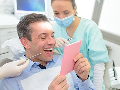 The image shows a man sitting in a dental chair with a smile on his face, holding up a pink cardboard sign with text, while a dentist wearing gloves and surgical mask stands behind him, observing the sign.