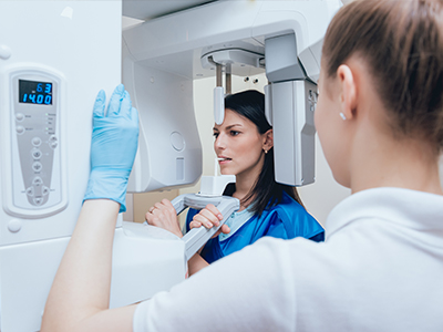 A woman stands beside a large medical imaging machine, possibly an MRI, while another person looks at her with interest. They are both wearing gloves, suggesting a sterile environment.