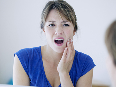 A woman with her mouth open, expressing concern or surprise, looking into a mirror while holding her hand on her chin.