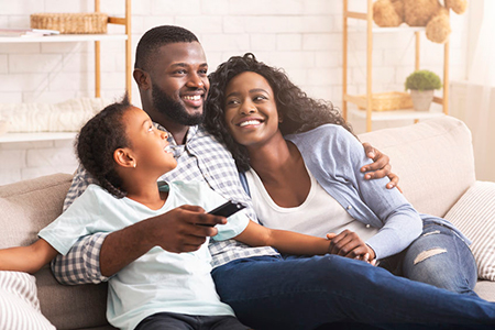 A family of four   a man, woman, and two children   sitting together on a couch, smiling at the camera, with the man holding a remote control.