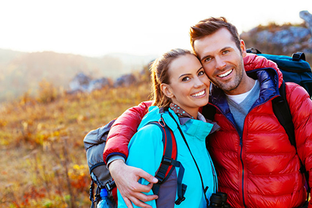 The image shows a man and woman posing together outdoors during daylight, with the man wearing a backpack and both dressed in casual hiking attire, against a scenic background of autumnal foliage and a clear sky.