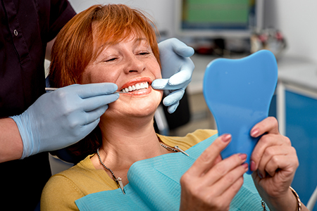 In the image, a woman with red hair is sitting in a dental chair, holding up a blue tooth model while a dentist works on her teeth.