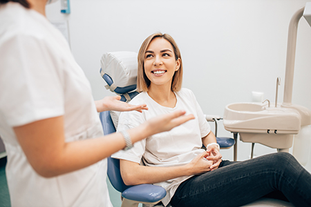This image depicts a woman sitting in a dental chair with a smile on her face, receiving dental care from a female dentist wearing white attire and holding a mirror.