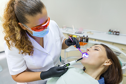 The image shows a dental hygienist performing a teeth cleaning procedure on a patient using an electric toothbrush while wearing personal protective equipment, including gloves and a mask.