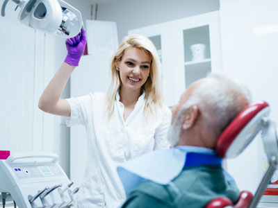 A dental hygienist in a white coat is showing a patient a dental device while standing next to a man sitting in a dental chair with his eyes closed, both are in a dental office setting.