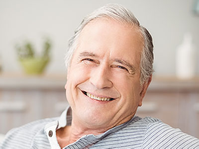 The image shows an elderly man with gray hair, smiling at the camera, wearing a blue shirt, sitting comfortably on a couch with his feet up.