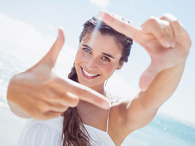 The image shows a woman with long hair, smiling at the camera, holding up her hand to frame a picture of herself against a sunny beach backdrop.